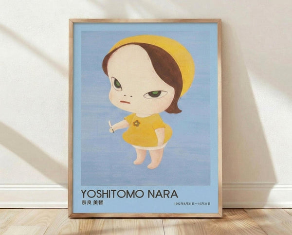 May include: Framed poster print leaning against white wall on light wooden floor. Shows stylized illustration of pale child figure with green eyes, brown bob haircut, yellow beret and dress against blue background. Text reads "Yoshitomo Nara" with Japanese characters and exhibition dates. Natural wood frame, soft lighting creates shadows on textured white wall.