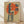 May include: Framed vintage-style poster leaning against white wall on wooden floor. Features woman in dark blue cheongsam dress sitting on wooden stool, sipping from small cup against large orange-red sun backdrop. Japanese characters on right side. Aged beige paper texture with distressed edges. Light wooden frame with natural wood grain visible.