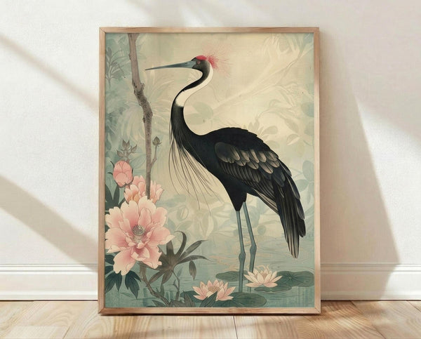 May include: Framed botanical art print featuring an elegant black crane with red head plume and long curved neck, standing among soft pink peonies and green foliage in vintage Japanese style. The artwork has muted sage green and cream tones, displayed in a natural wood frame against white textured wall on light wooden floor with soft window lighting.