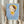 May include: Person holding framed poster featuring stylized cartoon girl character with brown hair, yellow beret and dress, holding small knife, against light blue background. Poster shows "YOSHITOMO NARA" text and Japanese characters. Person wears cream ruffled sleeves, holds dried wheat stems, neutral wall backdrop with soft window lighting.