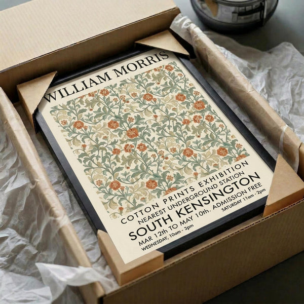 May include: Framed poster print featuring William Morris floral pattern in muted green, coral and cream tones displayed in cardboard shipping box with protective tissue paper wrapping and tape roll nearby on white fabric surface.
