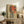 May include: Framed vintage-style poster featuring woman in dark blue cheongsam dress sitting on wooden stool against large red circular background with vertical Japanese characters, displayed on rustic wooden sideboard alongside fern plant, ceramic vase, and books in neutral-toned living room setting with beige armchair.