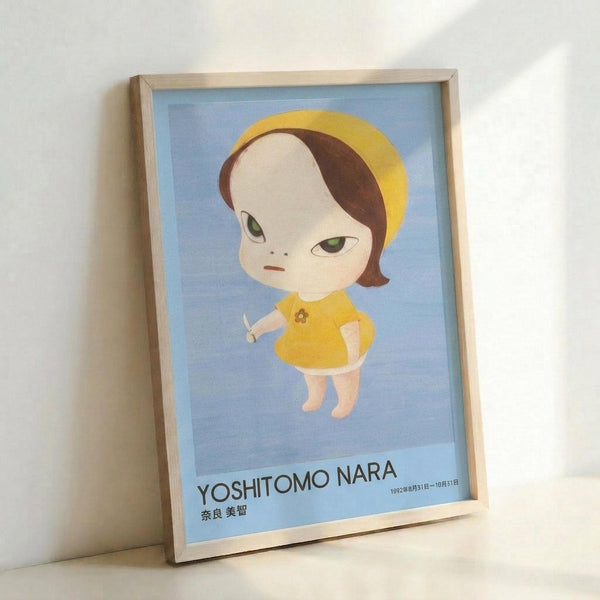Framed poster featuring a stylized illustration of a young girl with an oversized pale head, brown hair, and green eyes, wearing a yellow dress and holding a small knife. The character is rendered in a distinctive Japanese pop art style against a soft blue background. Text at bottom reads "YOSHITOMO NARA" with Japanese characters and exhibition dates. The poster is displayed in a light wooden frame leaning against a white wall.