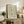May include: Framed William Morris exhibition poster with botanical floral pattern in sage green and coral colors, displayed on rustic wooden sideboard in cozy living room setting with fern plant, books, ceramic vases, and beige upholstered chair with brown leather cushion