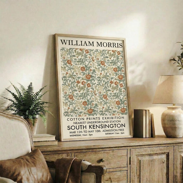 May include: Framed William Morris exhibition poster with botanical floral pattern in sage green and coral colors, displayed on rustic wooden sideboard in cozy living room setting with fern plant, books, ceramic vases, and beige upholstered chair with brown leather cushion