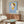 May include: Framed poster of stylized cartoon girl with brown hair, yellow dress and halo against blue background, displayed above wooden sideboard in modern living room with white curved chair, oval glass coffee table, and minimalist decor featuring warm wood tones and neutral colors.