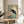 May include: A framed vintage-style artwork displayed on a round wooden side table beside a beige upholstered chair. The print features a black crane with red crown plumage standing among pink peonies and branches on a soft green background. Natural sunlight streams across the neutral-toned room setting with plant shadows visible on the white wall.