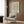 May include: Framed vintage floral poster with green foliage and orange flowers displayed on warm wood paneled wall above beige upholstered armchair with geometric patterned throw pillow. Modern brass floor lamp and round side table with open book complete the cozy reading nook scene with natural lighting from nearby window.