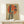 May include: Vintage-style poster in light wood frame showing woman in dark blue cheongsam dress sitting on stool against large red circle, with Chinese characters, displayed on white wooden mantel beside small white wrapped gift with evergreen sprig
