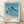 May include: Framed poster print leaning against white textured wall on light wooden floor. Artwork shows seven tall palm trees with long dark trunks against blue ocean and sky. Trees cast shadows on beige sand beach. Bottom features bold black text and row of colored dots in beige, light blue, teal, dark teal, and black. Light wood frame with white matting. Natural window light creates soft shadows on wall.