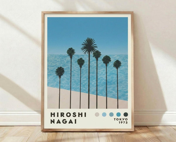 May include: Framed poster print leaning against white textured wall on light wooden floor. Artwork shows seven tall palm trees with long dark trunks against blue ocean and sky. Trees cast shadows on beige sand beach. Bottom features bold black text and row of colored dots in beige, light blue, teal, dark teal, and black. Light wood frame with white matting. Natural window light creates soft shadows on wall.