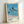 May include: Framed poster print leaning against white wall showing tropical beach scene with tall palm trees, blue ocean water, sandy beach, and clear sky. Features "HIROSHI NAGAI" text and "TOKYO 1973" with colored dots at bottom. Light wood frame with white matting creates clean, minimalist presentation in bright indoor setting.
