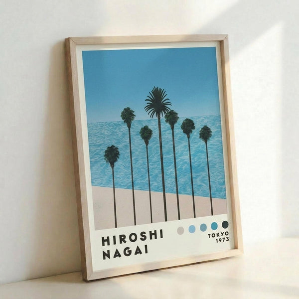 May include: Framed poster print leaning against white wall showing tropical beach scene with tall palm trees, blue ocean water, sandy beach, and clear sky. Features "HIROSHI NAGAI" text and "TOKYO 1973" with colored dots at bottom. Light wood frame with white matting creates clean, minimalist presentation in bright indoor setting.