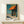 May include: Framed art print showing a coastal storefront with orange awning, teal blue sky, columns creating shadows on sidewalk, and lamp post. Displayed on light wood shelf with small wrapped gift box beside it against cream wall.