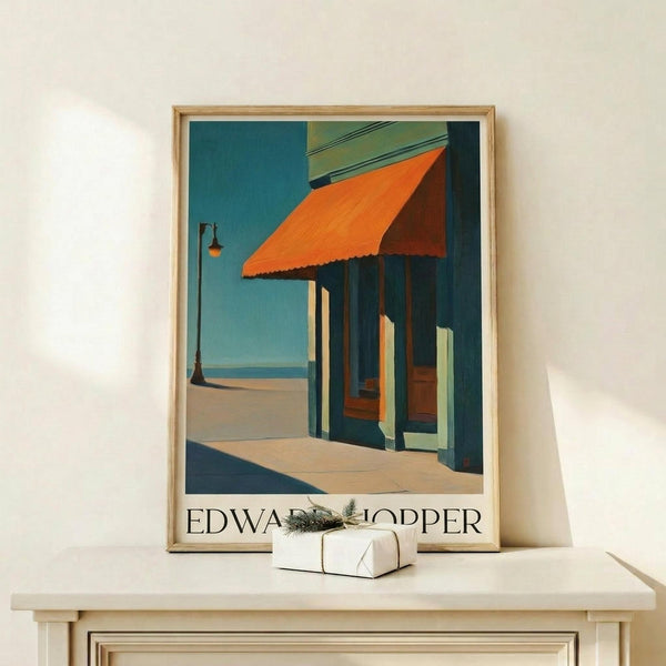 May include: Framed art print showing a coastal storefront with orange awning, teal blue sky, columns creating shadows on sidewalk, and lamp post. Displayed on light wood shelf with small wrapped gift box beside it against cream wall.