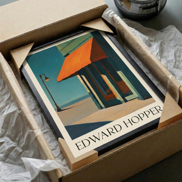 May include: Edward Hopper art print in cardboard box with protective wrapping. The poster shows a modernist building with orange triangular roof, teal walls, and large windows casting geometric shadows. Print appears medium-sized, nestled in brown packaging materials with rolled tubes nearby on white fabric surface.