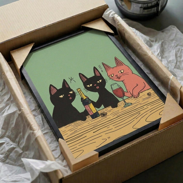 May include: Framed poster print in cardboard shipping box with protective wrapping. Artwork shows three stylized cats at wooden table - two black cats and one coral-pink cat sharing wine bottle and glass on sage green background. Simple illustration style with clean lines. Frame appears black, nestled in brown packaging materials.