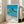 May include: Framed poster print leaning against white wall next to beige sofa in bright living room with large windows. Artwork shows row of tall palm trees silhouetted against bright blue sky and ocean with white sandy beach. Features minimalist design with five colored dots and "HIROSHI NAGAI" text at bottom, dated "TOKYO 1973". Natural wood frame, poster approximately 2-3 feet tall.