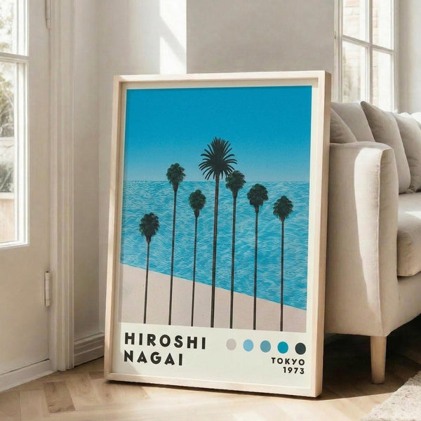 May include: Framed poster print leaning against white wall next to beige sofa in bright living room with large windows. Artwork shows row of tall palm trees silhouetted against bright blue sky and ocean with white sandy beach. Features minimalist design with five colored dots and "HIROSHI NAGAI" text at bottom, dated "TOKYO 1973". Natural wood frame, poster approximately 2-3 feet tall.