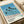 May include: Framed art print featuring stylized palm trees in silhouette against blue sky and ocean backdrop, displayed on light wooden surface next to beige fabric roll. The minimalist poster shows tall palm trees with long dark trunks and leafy crowns, rendered in muted tones of blue, beige, and green. Artist name and Tokyo 1973 text appear at bottom with small colored dots. Natural wood frame complements the coastal aesthetic.