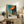 May include: Edward Hopper poster print in light wood frame displayed on rustic wooden sideboard in warm-toned living room. Artwork shows geometric architectural scene with orange awning, blue columns, and turquoise sky in Hopper's distinctive style. Styled with fern plant, stacked books, ceramic vessels, and cream upholstered furniture against white textured wall.