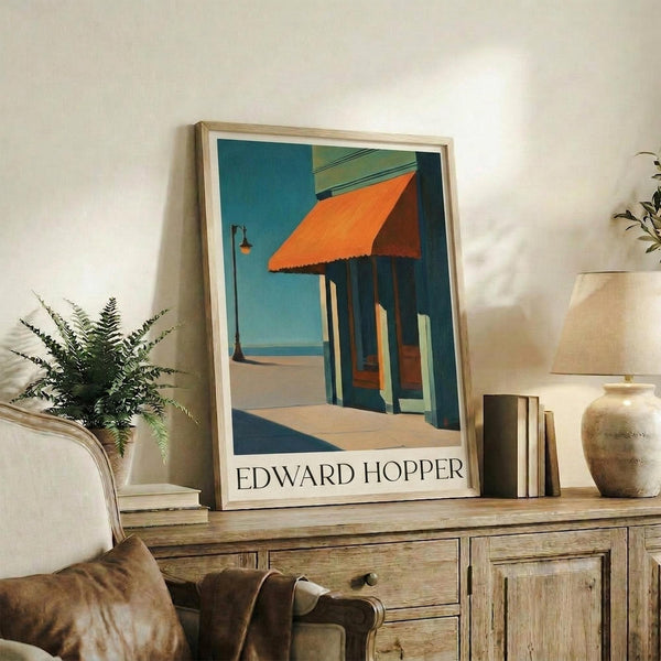 May include: Edward Hopper poster print in light wood frame displayed on rustic wooden sideboard in warm-toned living room. Artwork shows geometric architectural scene with orange awning, blue columns, and turquoise sky in Hopper's distinctive style. Styled with fern plant, stacked books, ceramic vessels, and cream upholstered furniture against white textured wall.