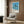 May include: Framed poster print displaying tropical beach scene with tall palm trees silhouetted against blue sky and ocean, mounted on wood-paneled wall above cream upholstered armchair with geometric patterned pillow, brass floor lamp and side table with open book nearby, showcasing mid-century modern interior styling with warm wood tones and neutral furnishings.