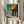 May include: Person in cream blouse holding framed Edward Hopper art print showing a storefront with bright orange awning, teal building facade, and clear blue sky. Light wooden frame against white curtained background with dried pampas grass stems visible on left side.