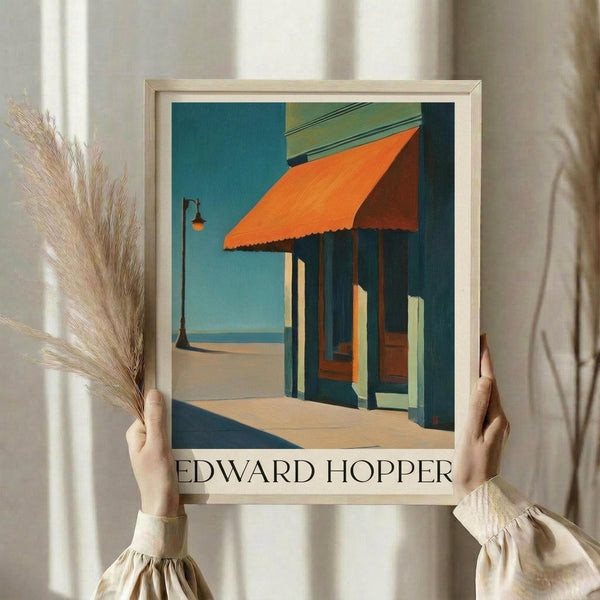 May include: Person in cream blouse holding framed Edward Hopper art print showing a storefront with bright orange awning, teal building facade, and clear blue sky. Light wooden frame against white curtained background with dried pampas grass stems visible on left side.