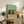 May include: Framed poster print with light wood frame showing three cartoon cats at wooden table - two black cats and one orange cat with wine bottle and glass on mint green background, displayed on rustic wooden console table alongside books, ceramic vase, and fern plant in cozy living room setting with beige chair visible.
