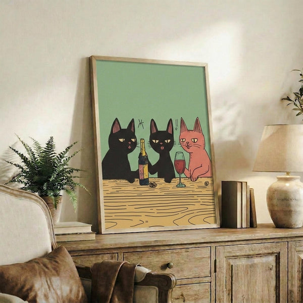 May include: Framed poster print with light wood frame showing three cartoon cats at wooden table - two black cats and one orange cat with wine bottle and glass on mint green background, displayed on rustic wooden console table alongside books, ceramic vase, and fern plant in cozy living room setting with beige chair visible.