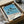 May include: Vintage-style poster print featuring tall palm trees silhouetted against blue sky and ocean, with beige sandy beach below. Art print shown in black frame inside brown cardboard packaging with protective wrapping, surrounded by white tissue paper on neutral surface.