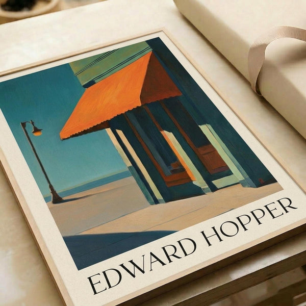 May include: Edward Hopper art print displayed on light wooden surface next to white ceramic vase. The artwork shows a mid-century modern house with distinctive orange triangular roof overhang, large windows, and teal-blue exterior walls. A street lamp stands in the foreground against pale blue sky. The print features Hopper's characteristic architectural style with clean geometric lines and bold color blocks in orange, teal, and cream tones.