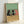 May include: Framed poster print leaning against white wall showing three stylized cats sitting at wooden table with wine bottle and glass. Two black cats and one orange cat drawn in simple cartoon style against sage green background. Light wood frame, casting soft shadow on white surface below.