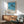 May include: Framed poster print displaying tropical palm trees silhouetted against bright blue ocean and sandy beach, with text reading "HIROSHI NAGAI TOKYO 1973" below the artwork. Shown hanging above a mid-century modern wooden credenza with table lamp, next to a mint green upholstered armchair with wooden frame. Natural sunlight creates shadows across white wall in minimalist room setting.