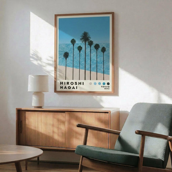 May include: Framed poster print displaying tropical palm trees silhouetted against bright blue ocean and sandy beach, with text reading "HIROSHI NAGAI TOKYO 1973" below the artwork. Shown hanging above a mid-century modern wooden credenza with table lamp, next to a mint green upholstered armchair with wooden frame. Natural sunlight creates shadows across white wall in minimalist room setting.