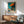 May include: Edward Hopper poster print in wooden frame hanging above mid-century modern sideboard with table lamp. Features painting of building with orange awning, teal blue walls and columns, beige ground. Room has green upholstered chair with wooden arms, round coffee table, white walls with dramatic sunlight shadows creating geometric patterns across the space.