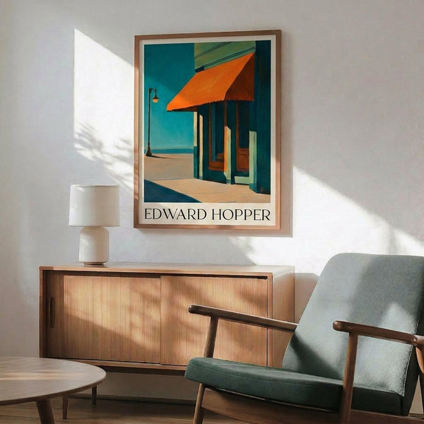 May include: Edward Hopper poster print in wooden frame hanging above mid-century modern sideboard with table lamp. Features painting of building with orange awning, teal blue walls and columns, beige ground. Room has green upholstered chair with wooden arms, round coffee table, white walls with dramatic sunlight shadows creating geometric patterns across the space.