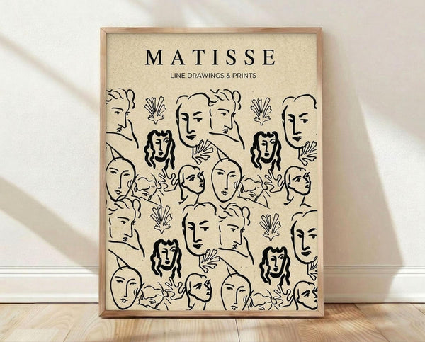 May include: A framed poster print featuring black line drawings of various human faces and leaf motifs in Matisse's distinctive style on cream-colored paper. The artwork shows repeating pattern of minimalist portrait sketches including women with long wavy hair, simplified facial features, and decorative botanical elements. Displayed in a light wood frame leaning against white walls on wooden flooring with soft natural lighting creating shadows.