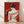 May include: Framed art print showing woman viewed from behind with dark hair in bun, holding red roses, wearing flowing white garment with red line details, set against deep red background. Natural wood frame displayed on light wooden floor against white wall with soft window light shadows.