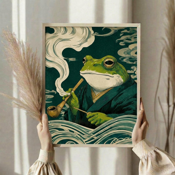 May include: Hands holding a framed art print featuring a green frog in traditional Japanese robes smoking a wooden pipe, rendered in ukiyo-e woodblock print style with teal and cream colors, stylized clouds and waves in background. Person wearing cream ruffled sleeves holds dried pampas grass stems. Light wooden frame against neutral curtained interior.