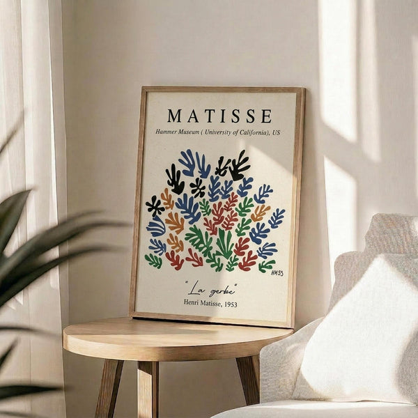 May include: Framed art print featuring colorful cut-out style botanical shapes in blue, green, orange, and red arranged in a circular pattern on cream background. Wood frame sits on round wooden side table next to beige upholstered chair with plant leaves visible in corner. Natural sunlight creates soft shadows on white wall behind.