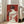 May include: Large framed poster with red background showing back view of woman with hair in bun holding roses, displayed in bright living room next to beige sofa. Artwork features vintage-style illustration in red and cream tones with light wood frame, leaning against white wall near windows.