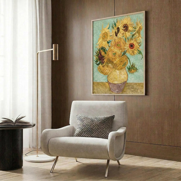 May include: Van Gogh's Sunflowers painting reproduction in thin gold frame hanging on wood paneled wall above cream upholstered armchair with geometric pillow. Bright yellow sunflowers in ceramic vase against turquoise background. Modern reading nook with brass floor lamp, side table with open book, and white curtains. Warm neutral interior with hardwood floors.