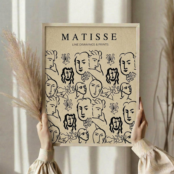 May include: Hands holding a framed beige poster featuring black line drawings of various faces and botanical elements in Matisse's minimalist style. The wooden frame sits against white curtains with dried pampas grass visible to the left. Person wears a cream-colored top with gathered sleeves.