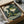 May include: A framed art print in a cardboard shipping box with protective packaging. The artwork shows a green frog character in traditional Japanese clothing smoking a wooden pipe, with white smoke swirls and stylized ocean waves in the background. The piece features muted green tones and classic ukiyo-e woodblock print styling. The black frame sits nestled in brown cardboard with white tissue paper padding.