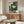 May include: Japanese-inspired artwork featuring a green frog in traditional robes smoking a pipe, surrounded by stylized waves and clouds in teal and cream colors. The print hangs above a mid-century modern walnut credenza in a minimalist living room with white walls, accompanied by a curved white chair and glass-top coffee table.