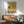 May include: Van Gogh's Sunflowers painting reproduction in wooden frame hanging above mid-century modern wooden sideboard with table lamp. Teal upholstered armchair with wooden arms sits nearby. Bright sunlight casts dramatic shadows across white wall. The artwork shows yellow sunflowers in ceramic vase against turquoise background.