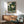 May include: Framed poster displaying a green frog character in traditional Japanese clothing smoking a wooden pipe, surrounded by stylized waves and clouds in green and cream tones. The artwork hangs above a wooden sideboard with a white ceramic lamp, next to a mid-century modern chair with sage green upholstery and wooden arms. Warm sunlight creates geometric shadows across the white wall and furniture in this contemporary living room setting.
