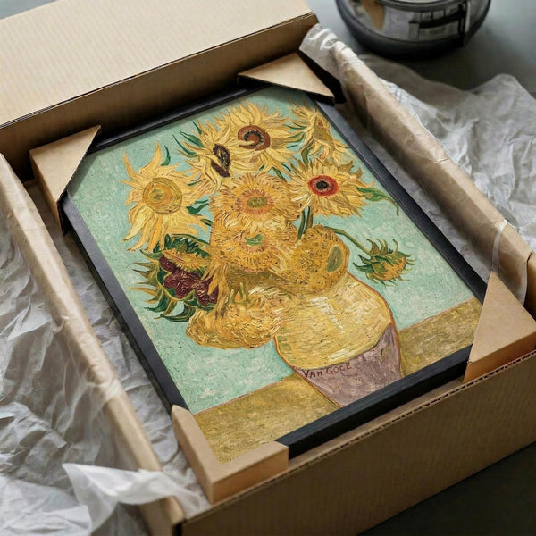 Framed reproduction of Van Gogh's sunflowers painting being unboxed from cardboard packaging with protective tissue paper. The artwork shows vibrant yellow sunflowers in a ceramic vase against a turquoise background, displayed in a black frame. Wooden packaging materials and bubble wrap are visible around the unboxing scene on a neutral surface.